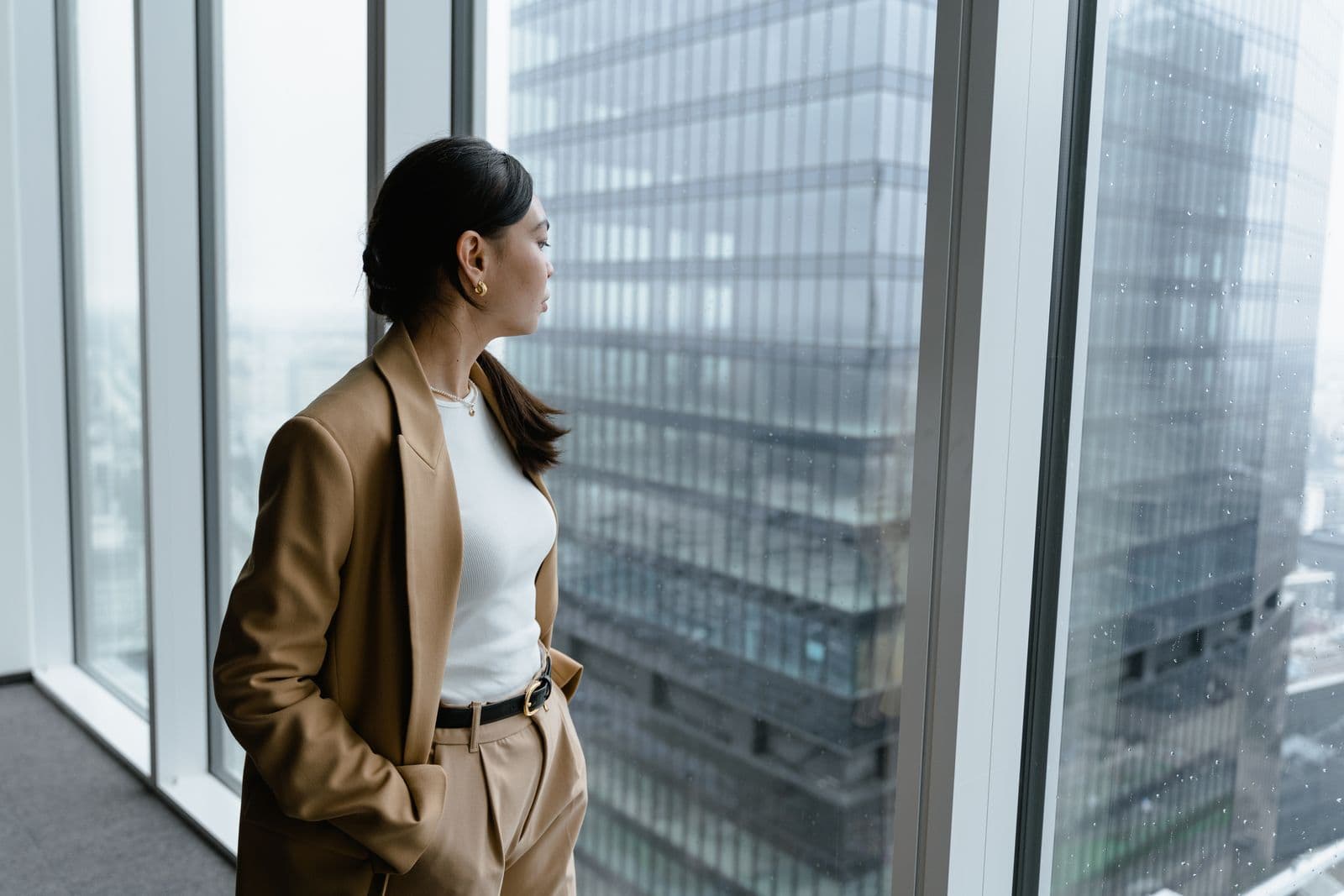 A woman in a camel blazer looking out a rain-flecked office window at glass towers